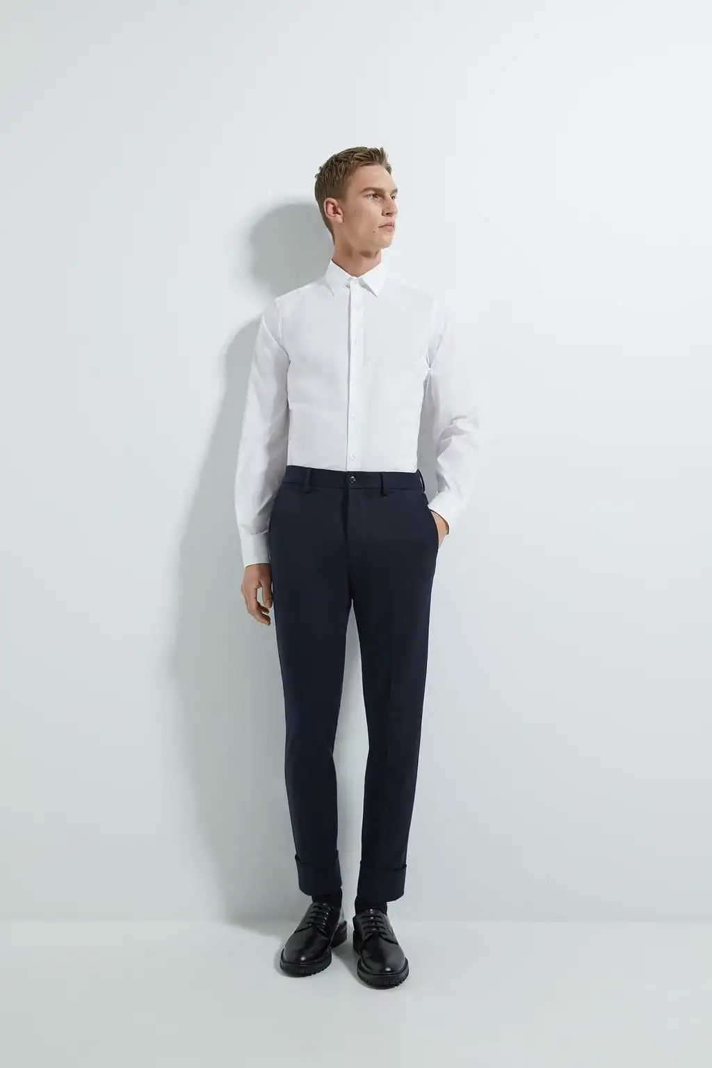 Stylish man wearing a crisp white long-sleeve dress shirt tucked into tailored navy trousers with black dress shoes, demonstrating modern formal menswear – Nordic Bespoke Tailor