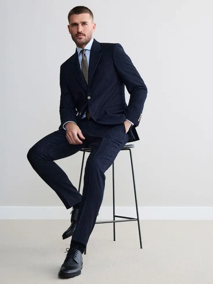 Male model sitting on stool wearing navy pinstripe suit with single-breasted blazer, matching trousers and brown tie – bespoke tailored menswear