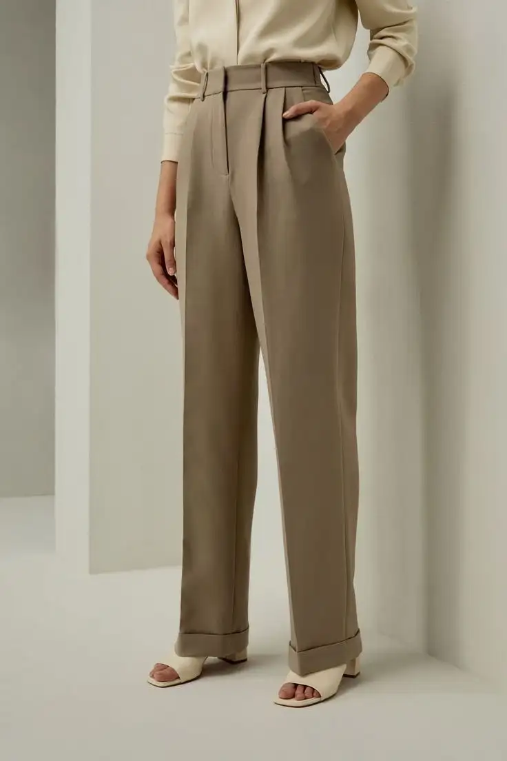 Female model wearing high-waisted beige pleated trousers with cuffed hems, paired with a tucked-in cream blouse and white sandals – bespoke women's tailored pants style
