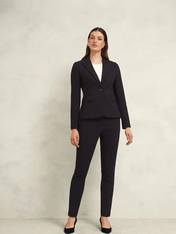 Woman wearing a fitted black blazer with a single button over a white top and matching slim-fit trousers, paired with black heels, embodying sleek and professional style.
