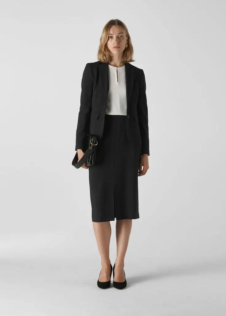 Chic woman wearing a black tailored skirt suit with a white blouse and black pumps, holding a small clutch bag, exemplifying refined professional attire