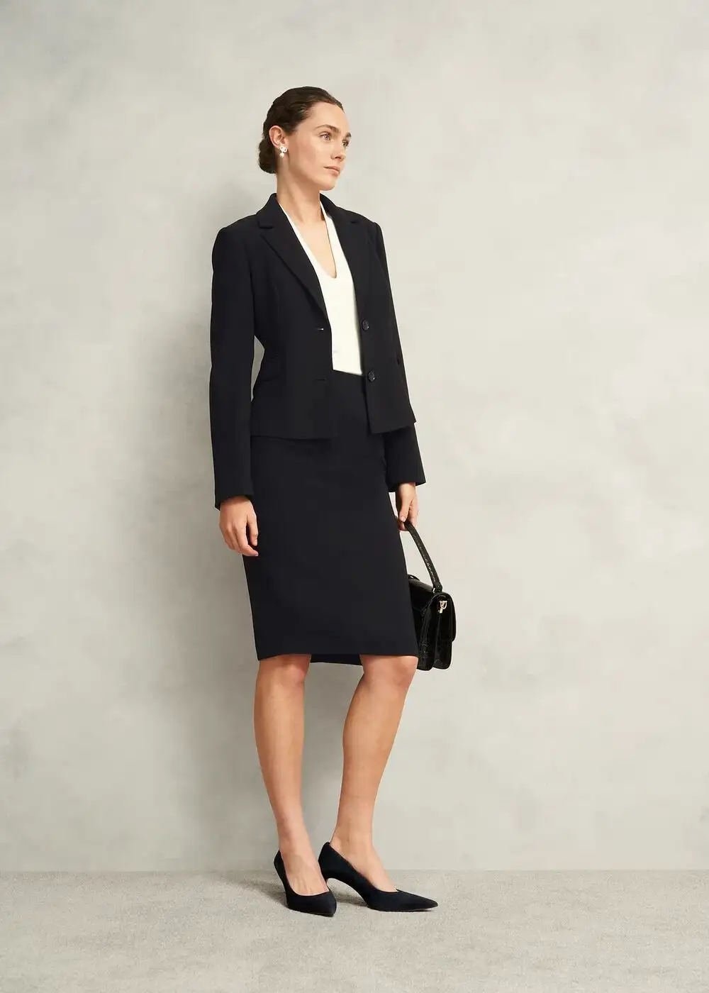 Professional woman wearing a navy skirt suit with a tailored blazer and knee-length skirt, paired with a white blouse, black heels and a handbag, conveying polished office attire