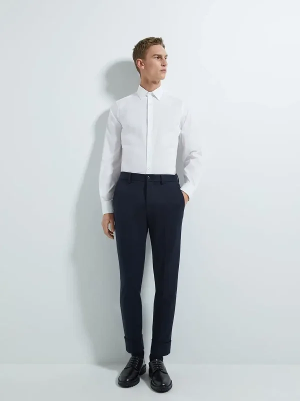 Stylish man wearing a crisp white long-sleeve dress shirt tucked into tailored navy trousers with black dress shoes, demonstrating modern formal menswear – Nordic Bespoke Tailor