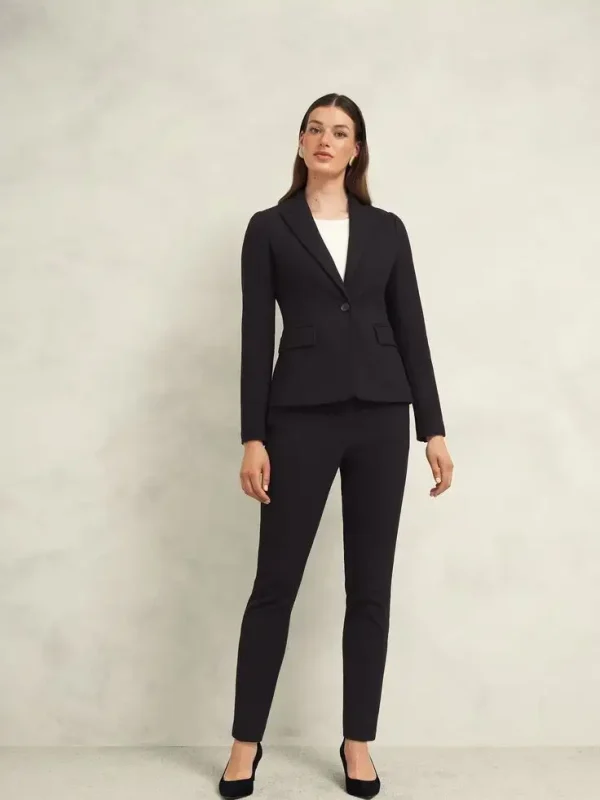Woman wearing a fitted black blazer with a single button over a white top and matching slim-fit trousers, paired with black heels, embodying sleek and professional style.
